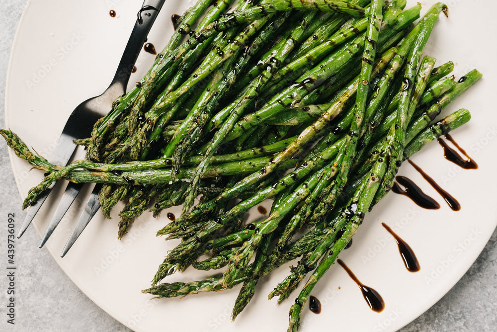 Steamed asparagus on a white plate with a drizzle of balsamic vinegar, on a gray surface.