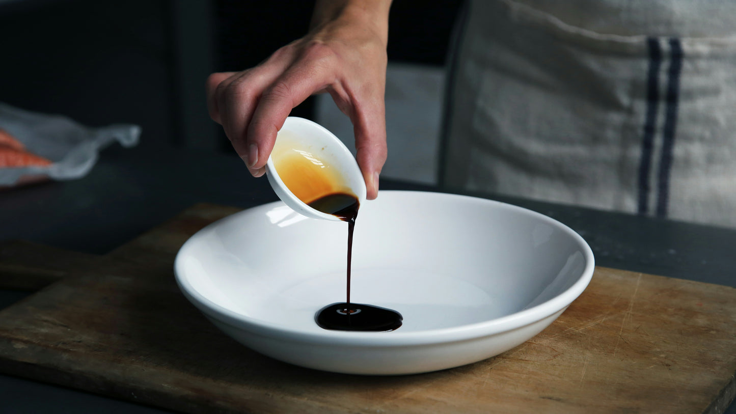 Hand pouring balsamic vinegar from a small white container into a larger white bowl on a wooden surface.