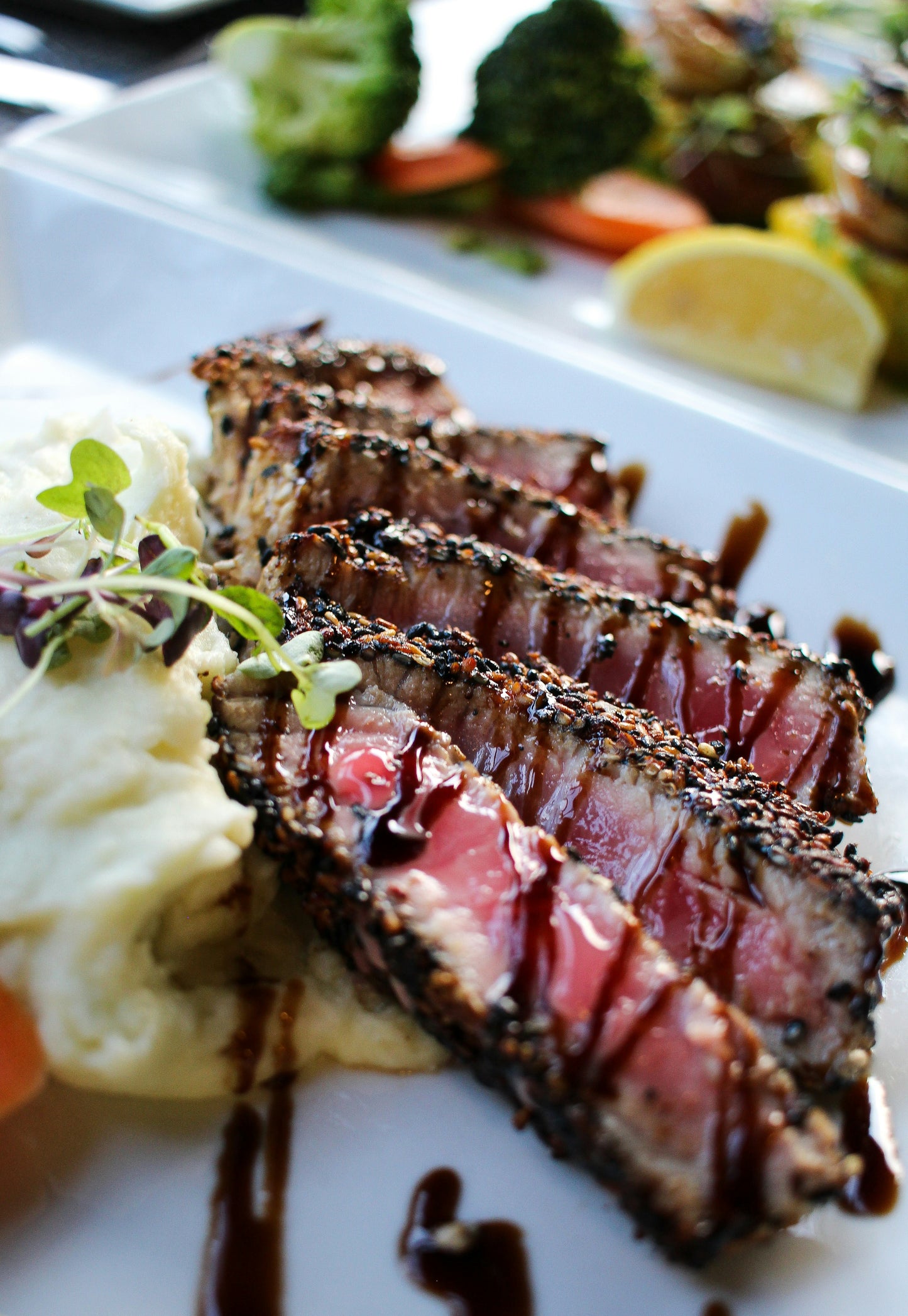 Sliced seared steak drizzled with balsamic vinegar with a side of mashed potatoes and vegetables on a white plate.