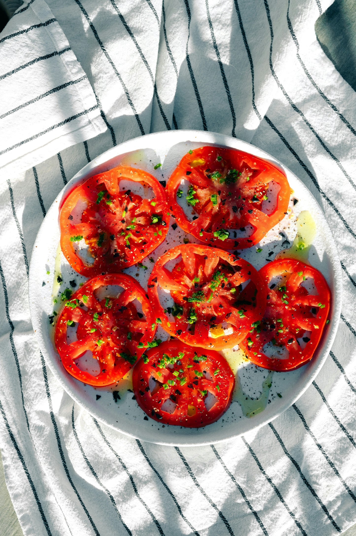 Sliced tomatoes on a white plate with herbs, placed on a striped fabric background