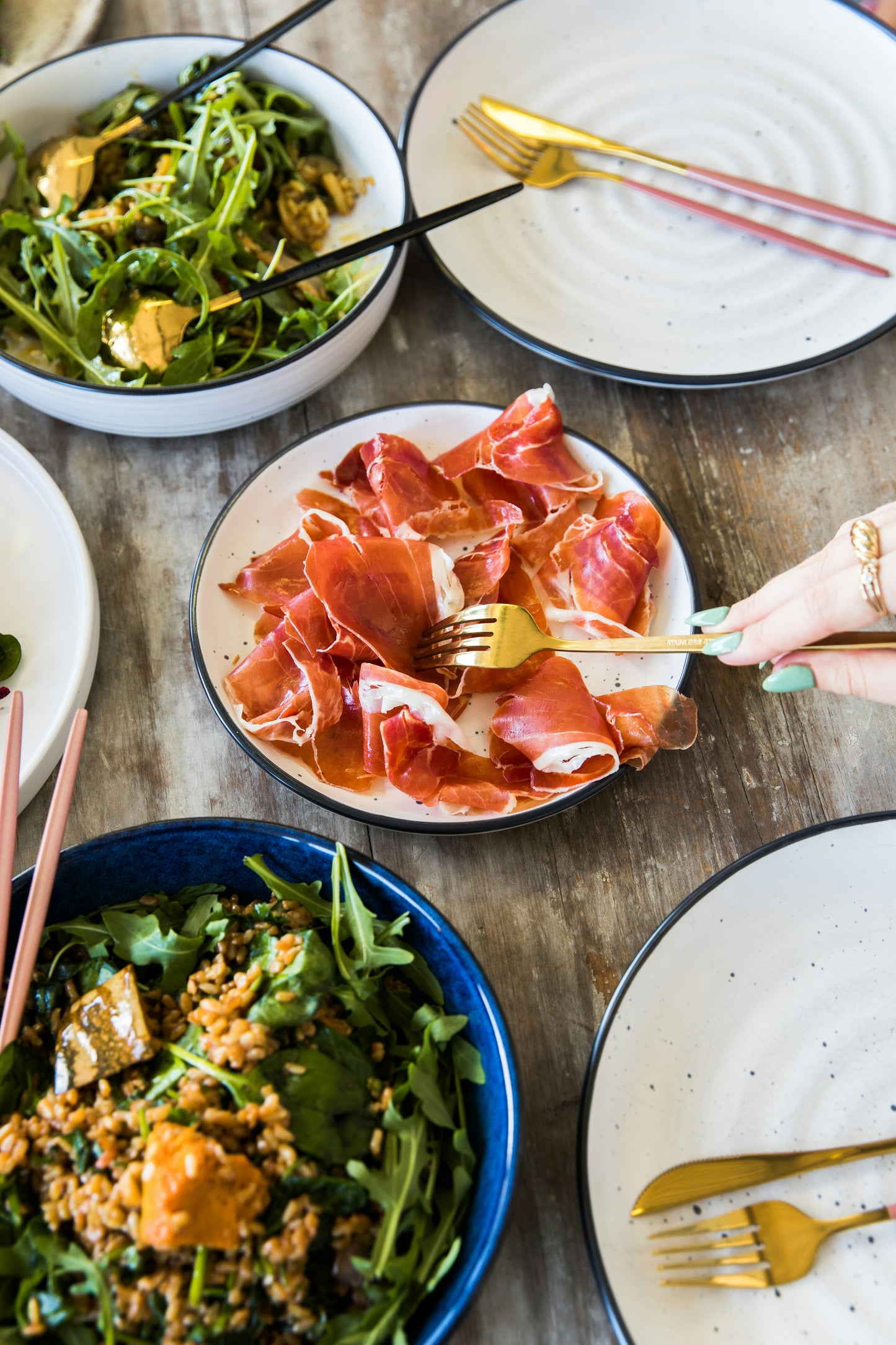 Dinner table with plates of food including arugula and prosciutto, and a hand holding a fork.