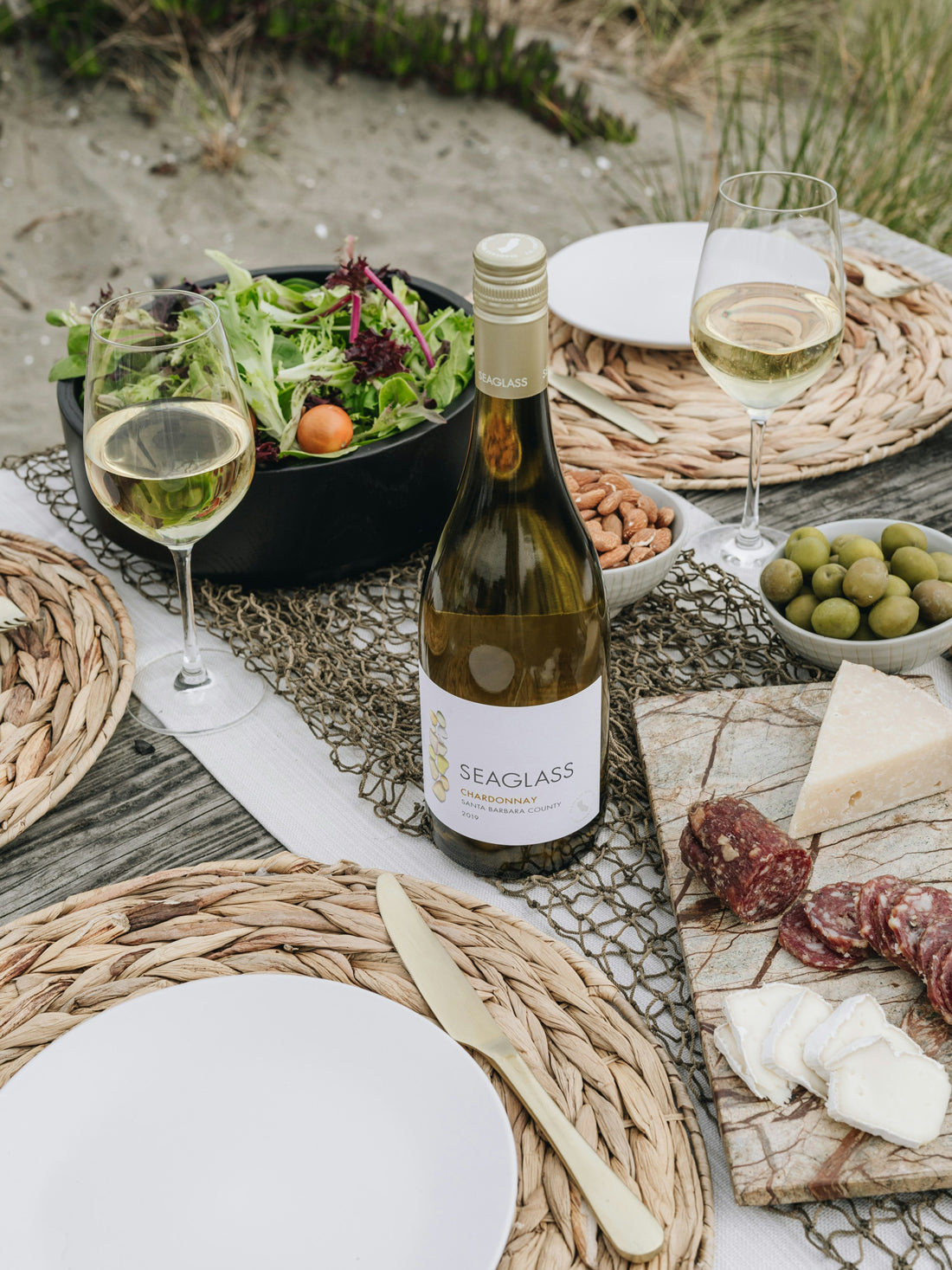Outdoor picnic setup with wine, cheese, and salad on a sandy beach.
